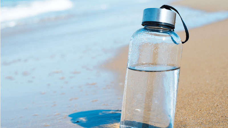 A reusable water bottle with water in it sits in the sand at the beach near the waters edge