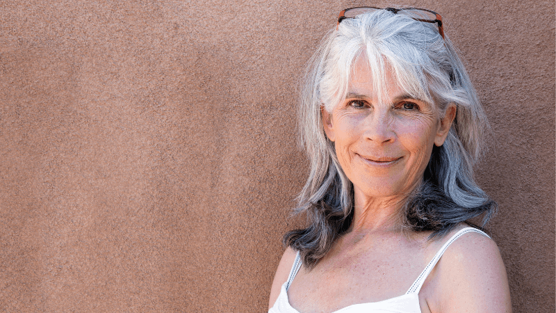 A woman with white and grey hair smiles while standing against a brown wall