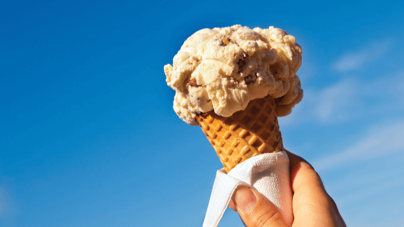 A scoop of ice cream in a cone is held up to a blue sky