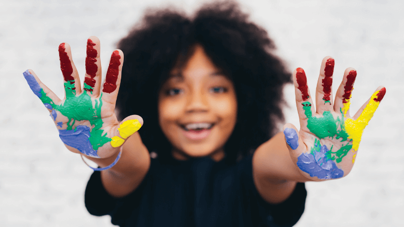 A child with paint on their hands reaches towards the camera