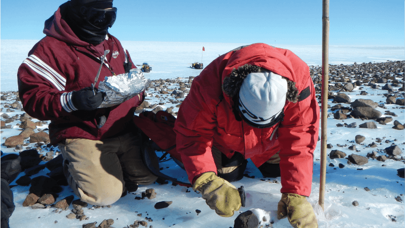 Two scientists in red winter jackets dig in ice around a rock