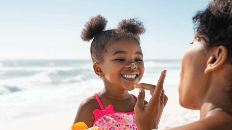 A young child on the beach smiles while someone applies sunscreen to her face