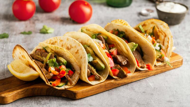 A wooden board with soft shelled tacos on it.