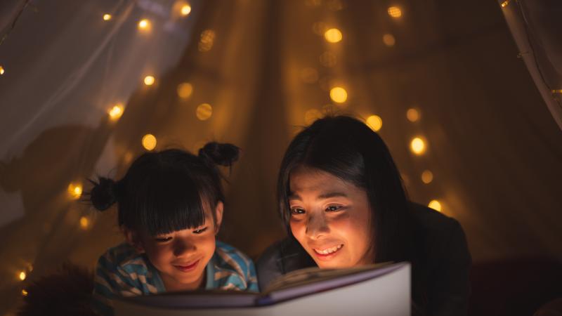 A young Asian mother reads a story from a book to her daughter while sitting on the bed at home.