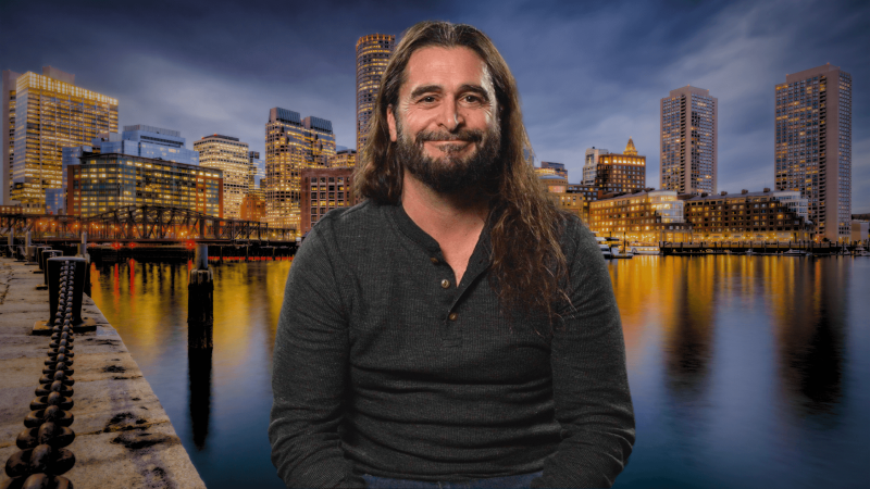 Drew Rollert in front of a harbor side view of Boston