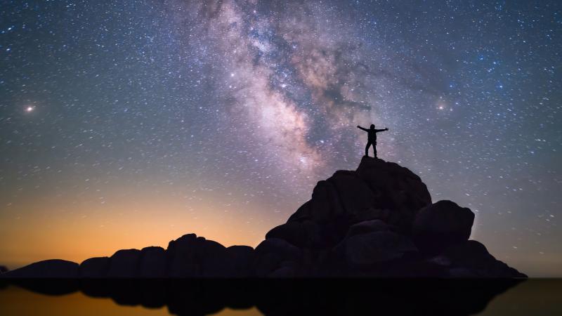 A person on a tall rock with arms outstretched, looking up at the night sky