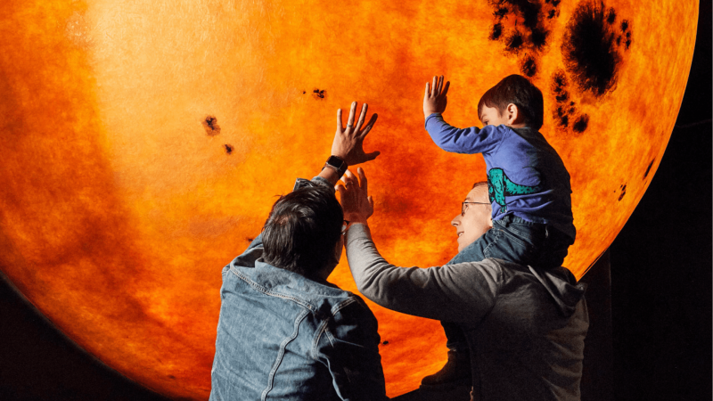 Two adults and one young visitor touch a model sun in the Museum of Science.