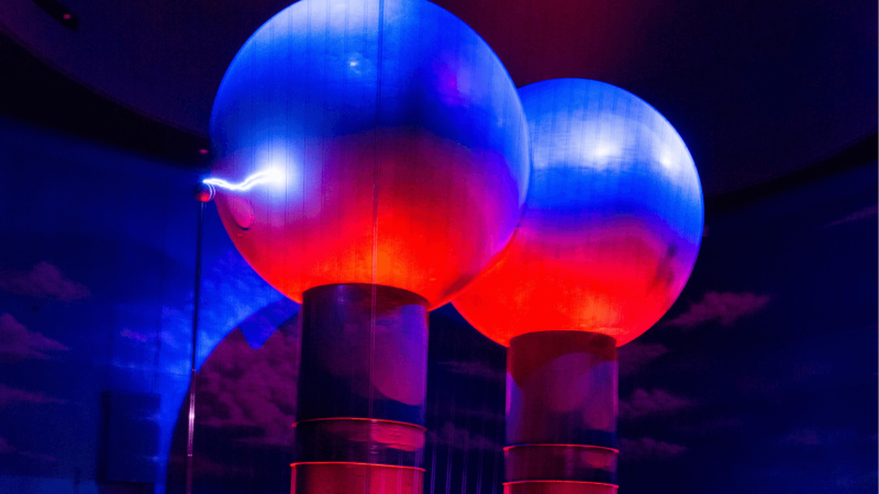 A moment in the Theater of Electricity showing the Van der Graaf generator lit in red and blue lights with a lightning bolt coming out the side.