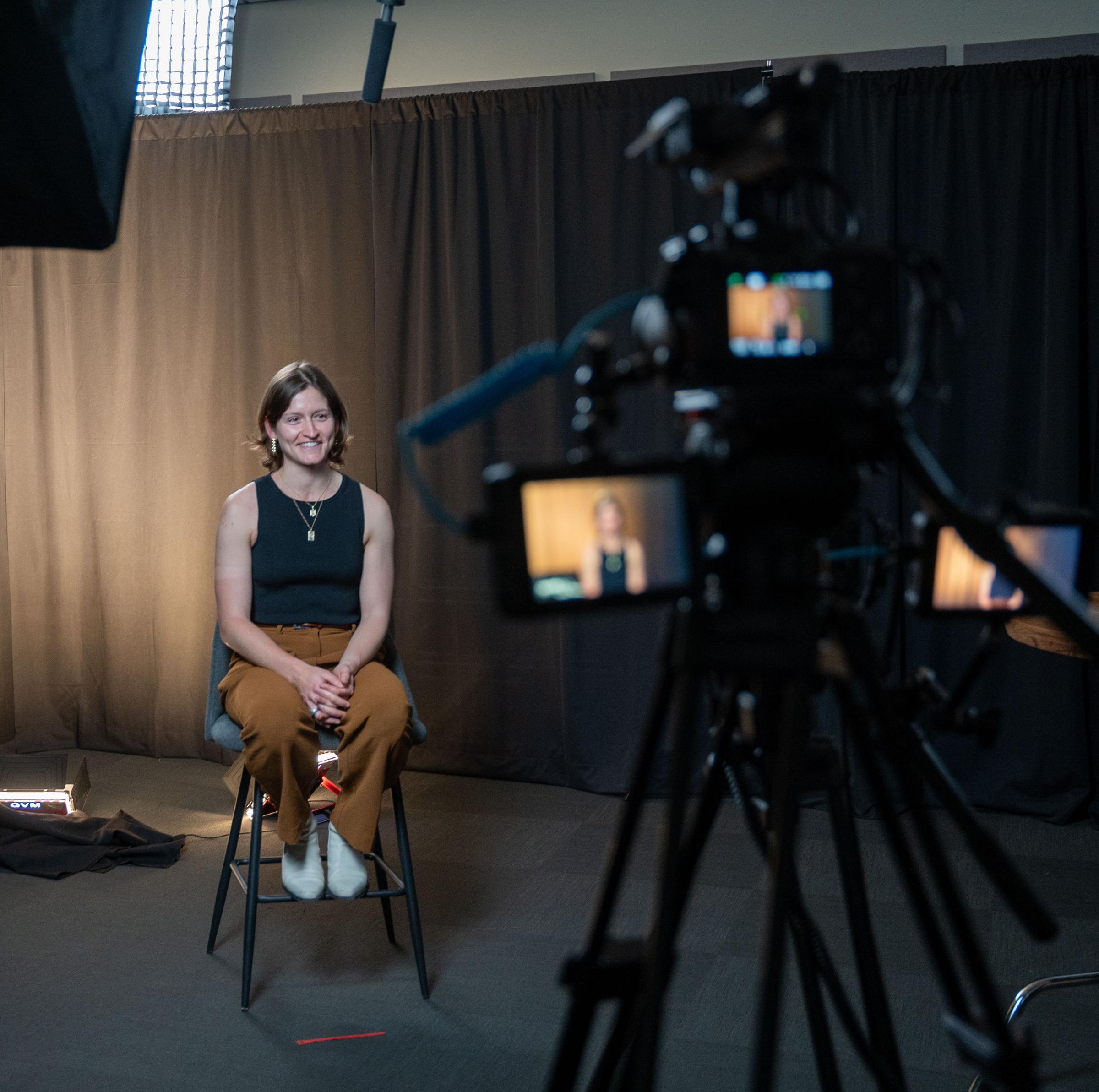 A woman in a black top and khaki pants smiles at a camera with cameras and lights in the view.
