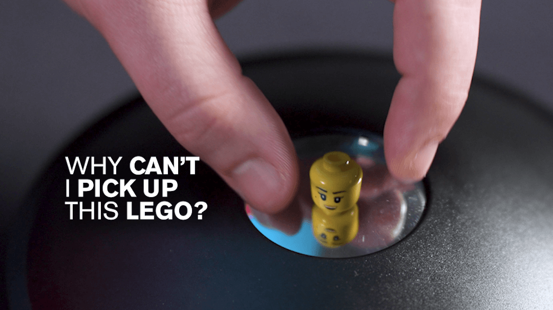 An image of a Lego head reflected in parabolic mirrors with white text reading "Why can't I pick up this Lego?"