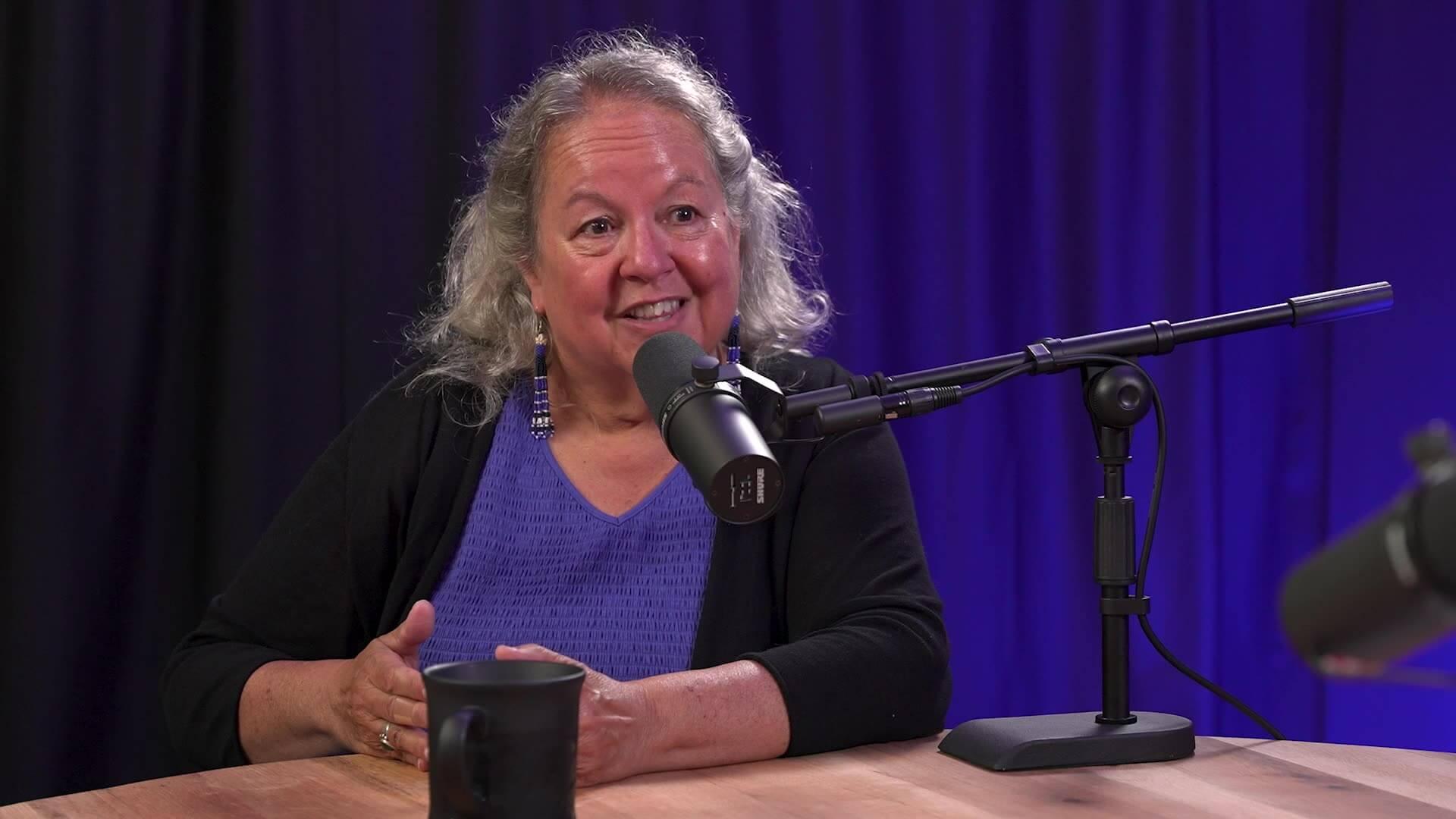 Robin Wall Kimmerer talking into a microphone in front of a purple lit curtain