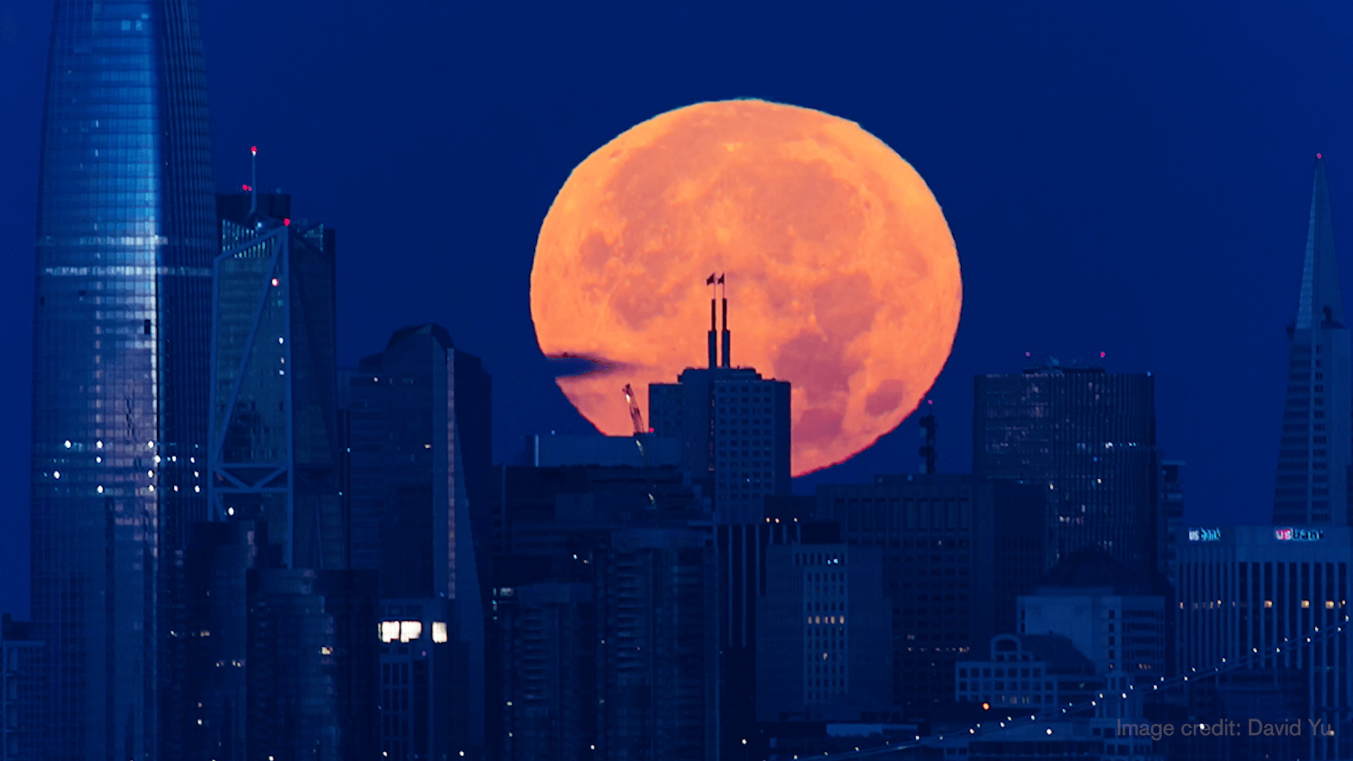 A large moon rising behind a city skyscraper.