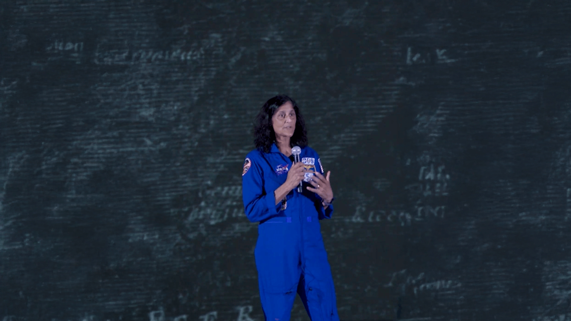 Suni Williams in a blue NASA jumpsuit in front of a chalkboard wall.