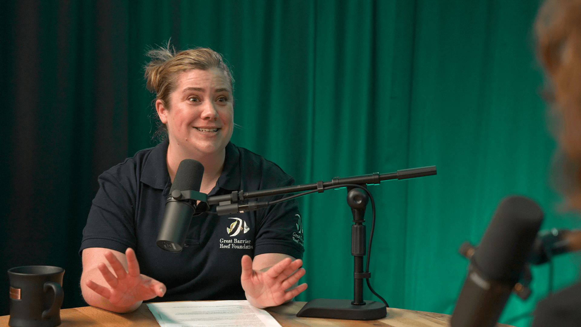 Jane Adcroft smiles while speaking into a microphone in front of a curtain lit with green light