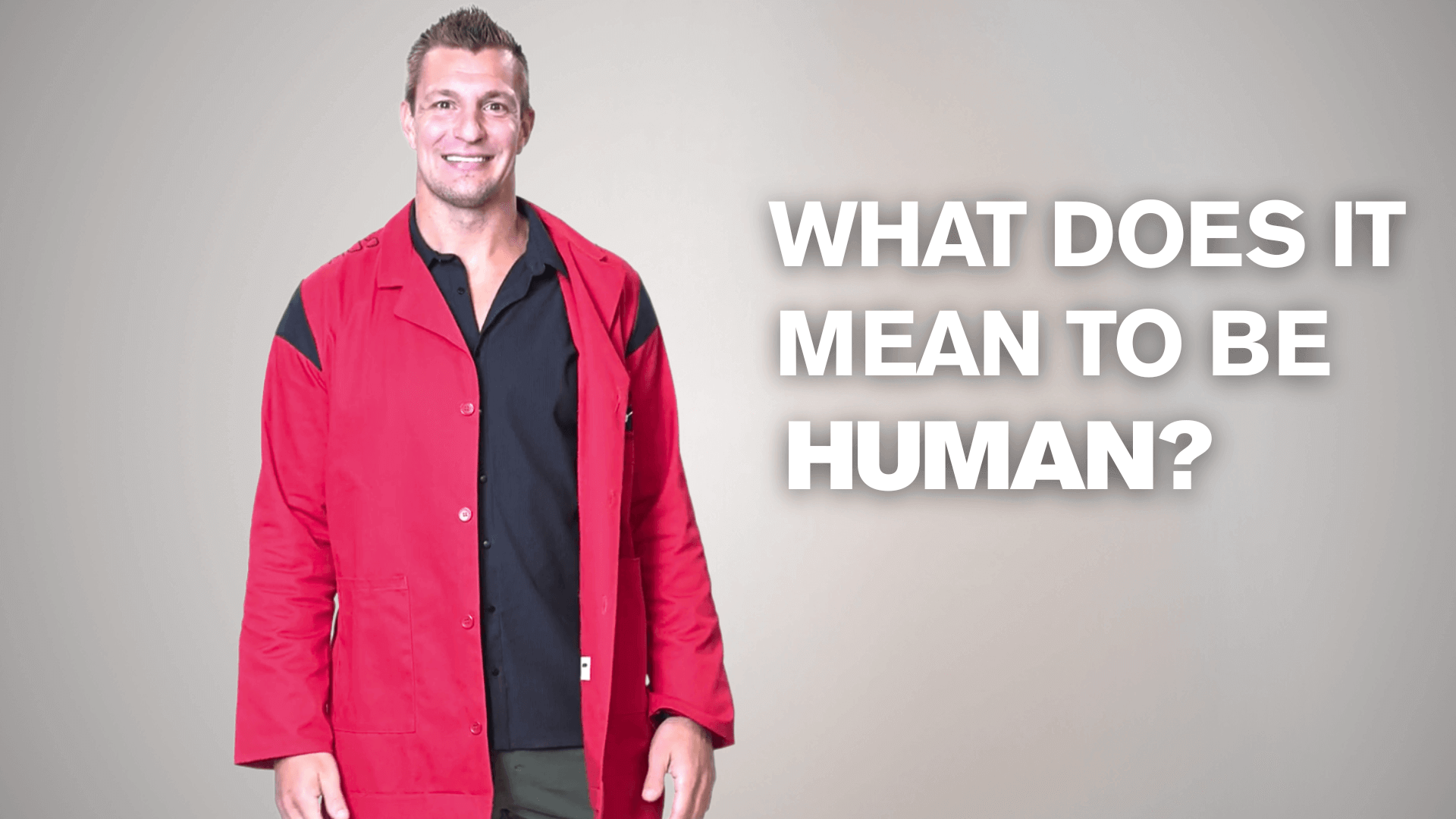 Rob Gronkowski smiles at the camera while wearing a red lab coat with white text reading "What does it mean to be human?"