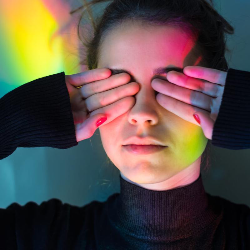 A figure holding hand, palms forward, over their eyes. Lit by various colored lights.